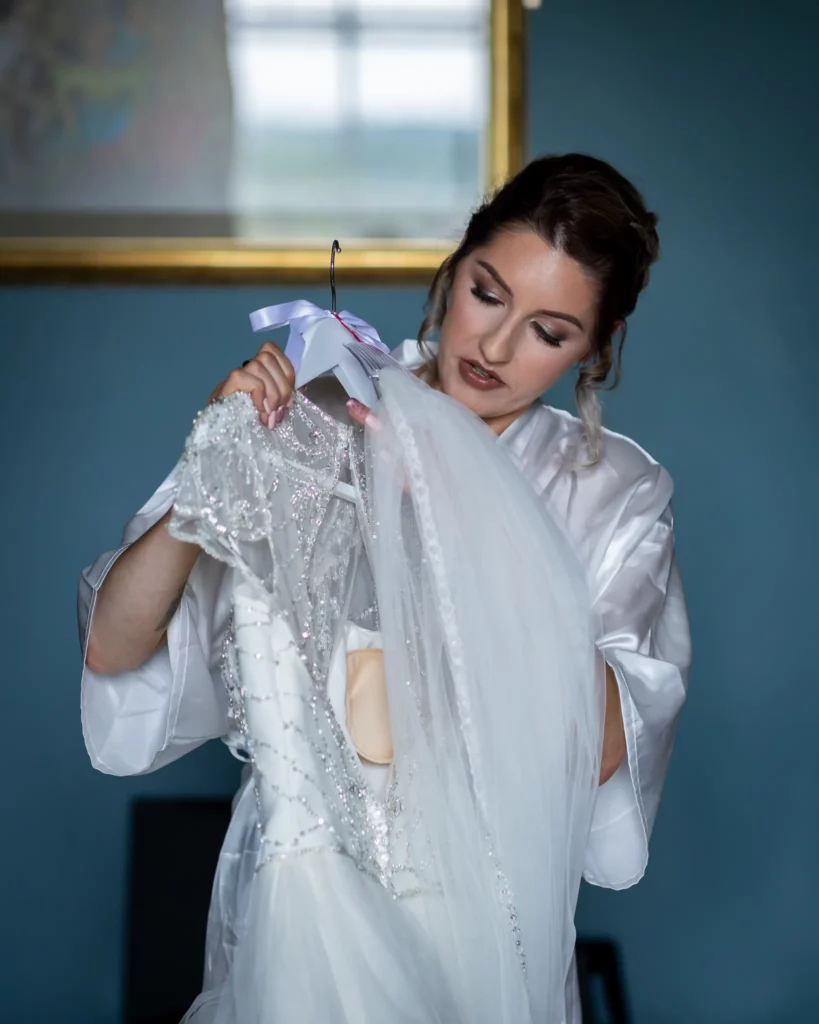 A woman in a white dressing gown holds and examines a white wedding dress on a hanger in a dimly lit room. Copyright Somerside Photography Ltd // www.somersidephotography.com