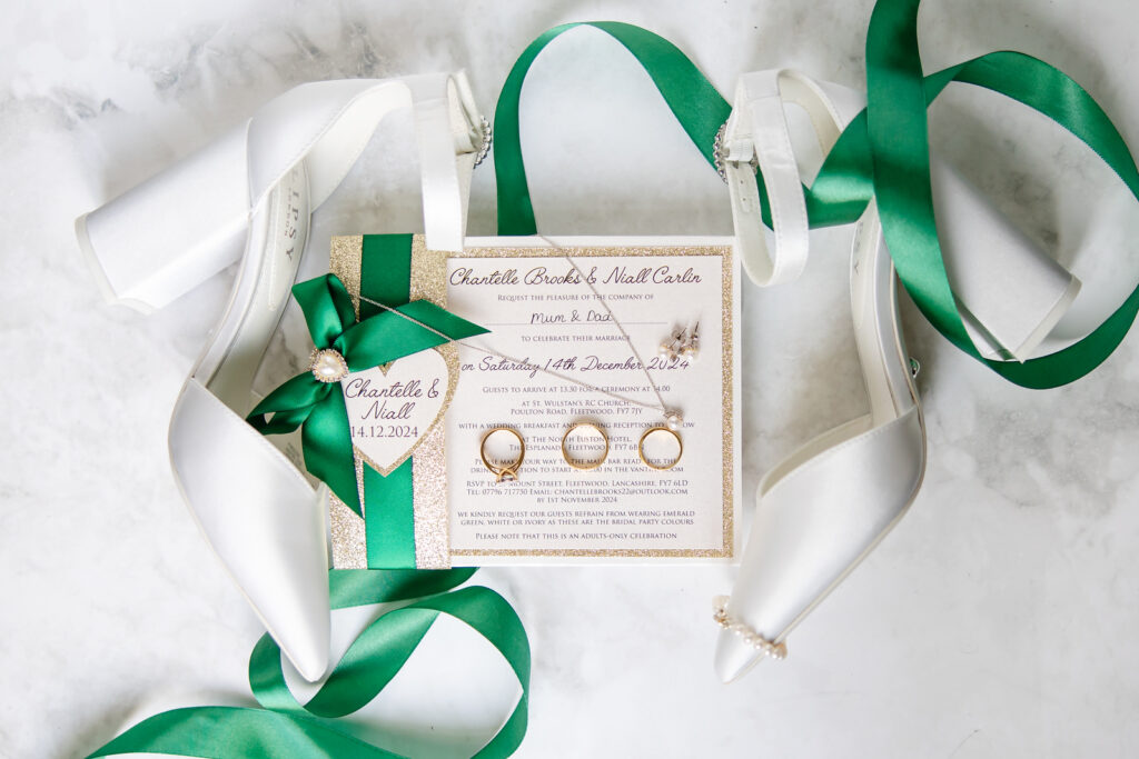 Flat lay of a wedding invitation, three gold rings, white high-heeled shoes with green ribbons, and a green ribbon with a pearl and date charm on a white marble surface. Copyright Somerside Photography Ltd // www.somersidephotography.com