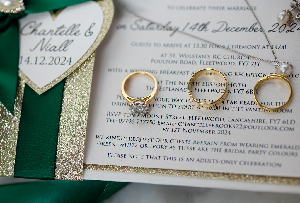 Three gold rings are placed on a wedding invitation card next to a green and gold heart-shaped decoration labelled "Chantelle & Niall 14.12.2024. Copyright Somerside Photography Ltd // www.somersidephotography.com
