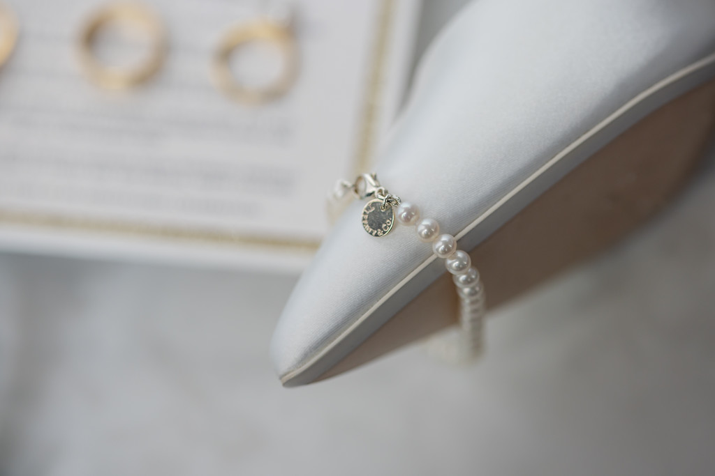 A close-up of a white satin high-heeled shoe with a pearl anklet and small gold charm draped over the heel. Copyright Somerside Photography Ltd // www.somersidephotography.com