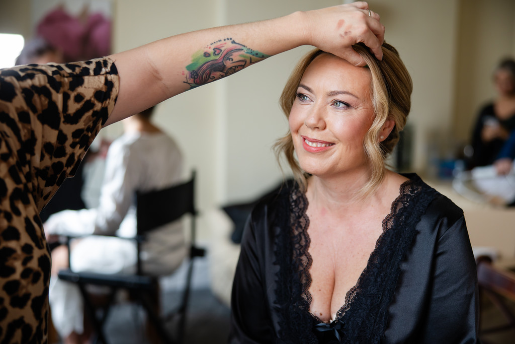 A woman in a black robe smiles whilst a make-up artist applies make-up to her forehead; others are visible in the blurred background. Copyright Somerside Photography Ltd // www.somersidephotography.com