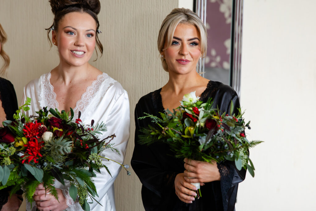 Two women in white and black robes hold bouquets of red and green flowers, standing indoors against a neutral wall. Copyright Somerside Photography Ltd // www.somersidephotography.com