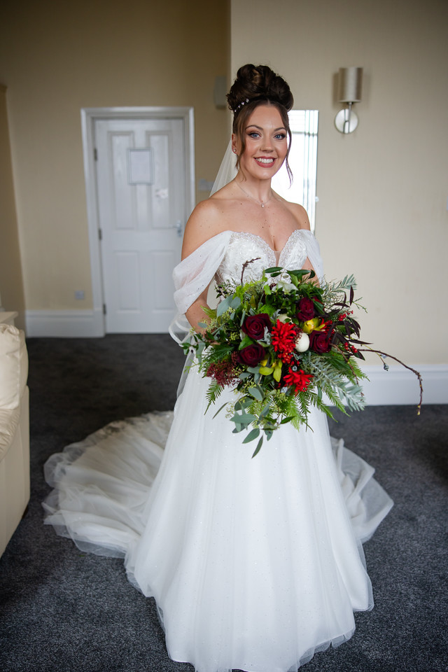 A bride in a white wedding dress and veil stands indoors holding a bouquet of red and green flowers, smiling at the camera. Copyright Somerside Photography Ltd // www.somersidephotography.com