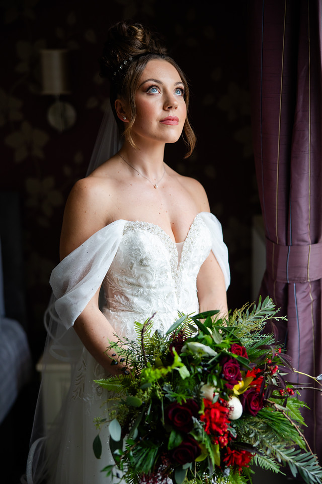 A bride in an off-the-shoulder white gown holds a bouquet of red and green flowers, standing by a window with natural light illuminating her face. Copyright Somerside Photography Ltd // www.somersidephotography.com