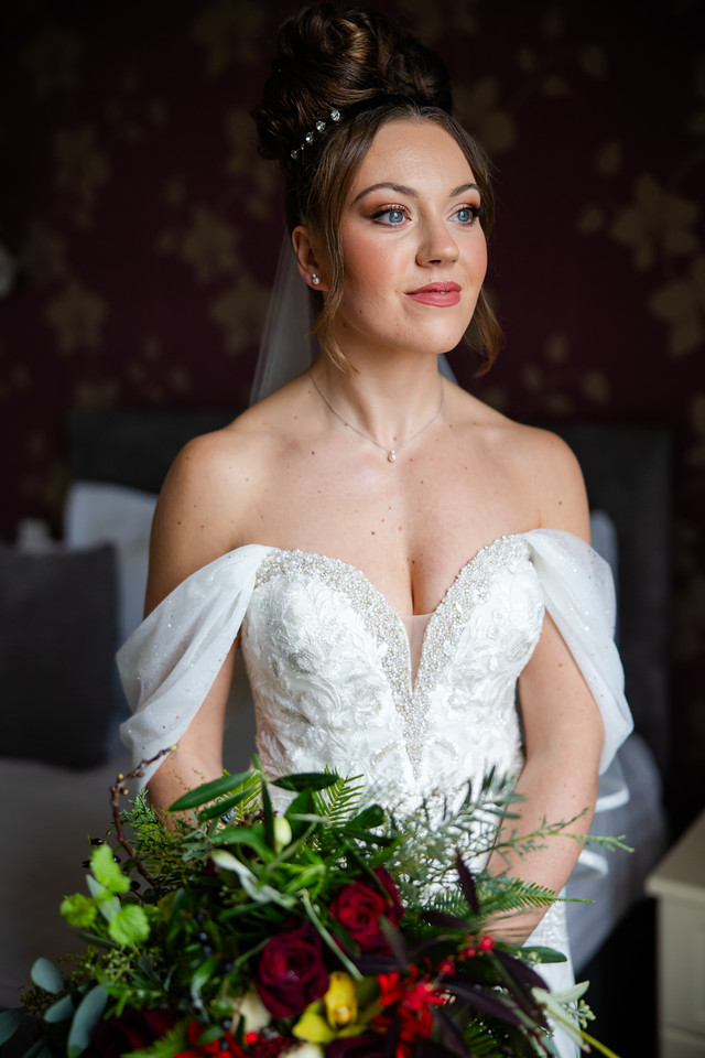 A woman in a white, off-the-shoulder wedding dress holds a bunch of red and green flowers, standing indoors against a blurred background. Copyright Somerside Photography Ltd // www.somersidephotography.com