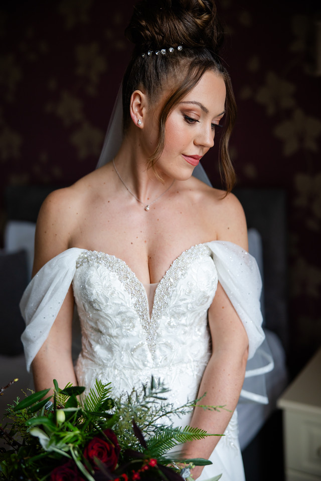 A bride in a white, off-the-shoulder wedding dress holds a bunch of red and green flowers, looking down with a neutral expression. Copyright Somerside Photography Ltd // www.somersidephotography.com
