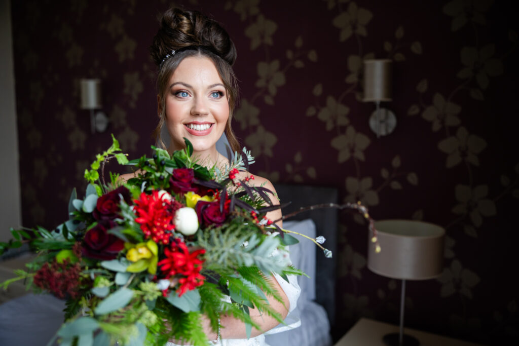 A woman in a white dress holds a large bouquet of flowers, standing indoors in a room with floral-patterned wallpaper and a bed in the background. Copyright Somerside Photography Ltd // www.somersidephotography.com