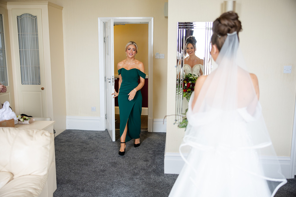 A woman in a green dress enters a room, smiling and looking at a bride in a white wedding dress who stands with her back to the camera. Copyright Somerside Photography Ltd // www.somersidephotography.com
