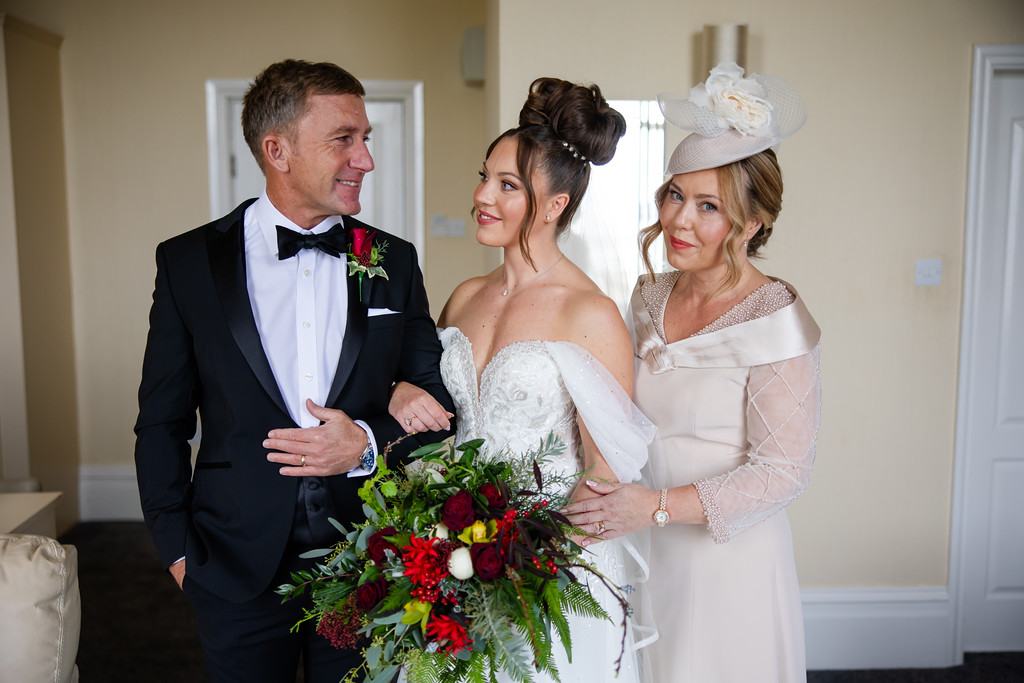 A bride in a white dress holds a bouquet, standing between a man in a tuxedo and a woman in a cream outfit and hat. All three are smiling indoors. Copyright Somerside Photography Ltd // www.somersidephotography.com
