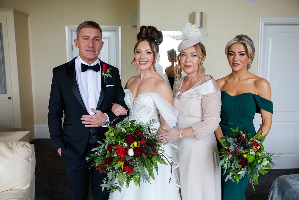 Four people dressed in formal attire pose indoors, including a bride holding a bouquet, a man in a dinner suit, and two women in elegant dresses with floral posies. Copyright Somerside Photography Ltd // www.somersidephotography.com