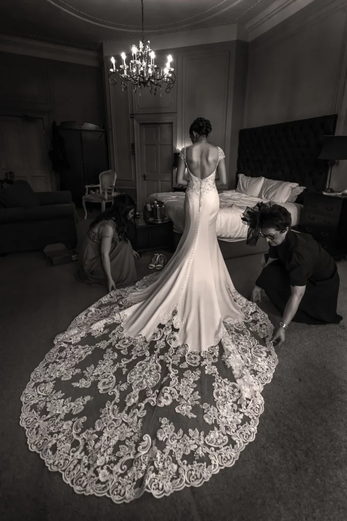 A bride stands in a room wearing a fitted wedding dress with a long lace train as two women adjust the dress. Copyright Somerside Photography Ltd // www.somersidephotography.com