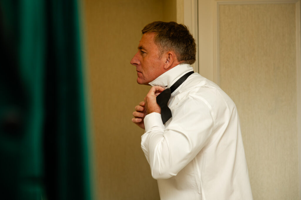 A man in a white dress shirt is standing indoors, adjusting a black tie around his collar near a cream-coloured wall. Copyright Somerside Photography Ltd // www.somersidephotography.com