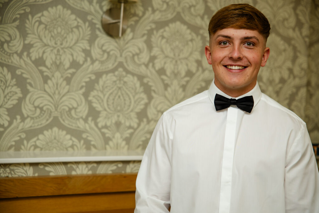 A young man wearing a white dress shirt and black bow tie stands smiling in front of a patterned wallpaper background. Copyright Somerside Photography Ltd // www.somersidephotography.com