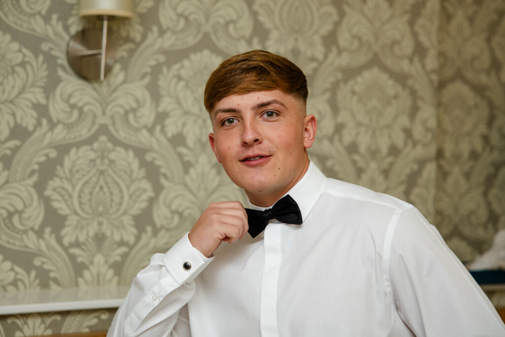 A young man in a white dress shirt adjusts a black bow tie, standing in front of patterned wallpaper. Copyright Somerside Photography Ltd // www.somersidephotography.com