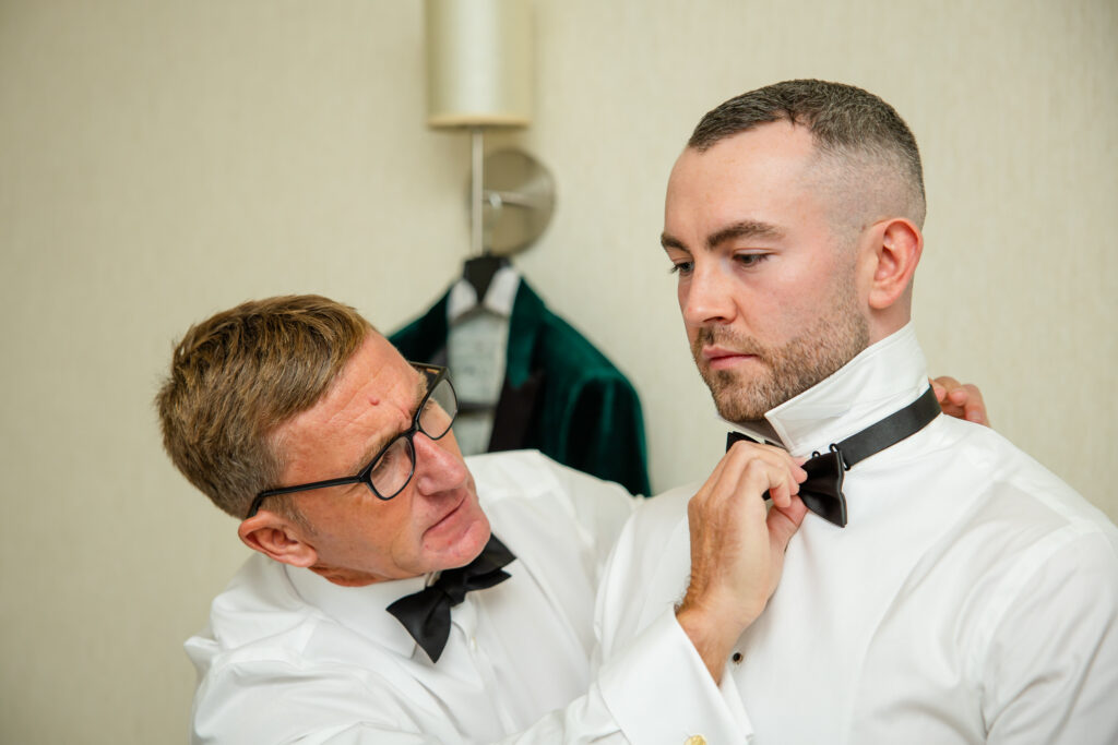 Two men in formal white shirts and bow ties; one helps the other adjust his collar in a room with a suit jacket hanging in the background. Copyright Somerside Photography Ltd // www.somersidephotography.com