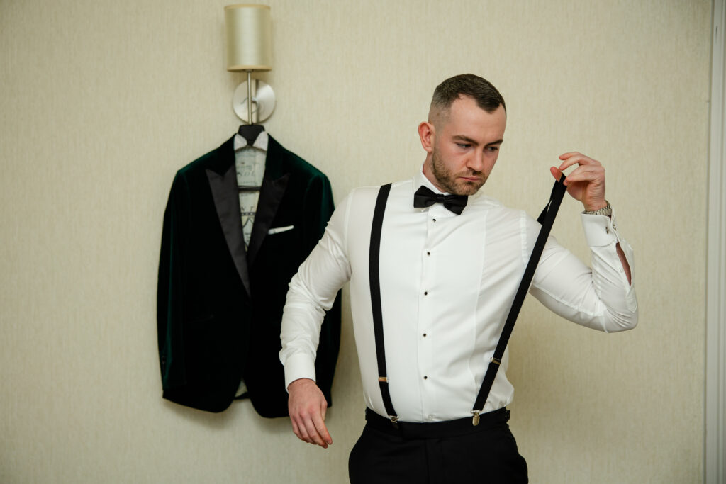 A man in formal attire adjusts his black braces; a dark blazer hangs on the wall behind him. Copyright Somerside Photography Ltd // www.somersidephotography.com