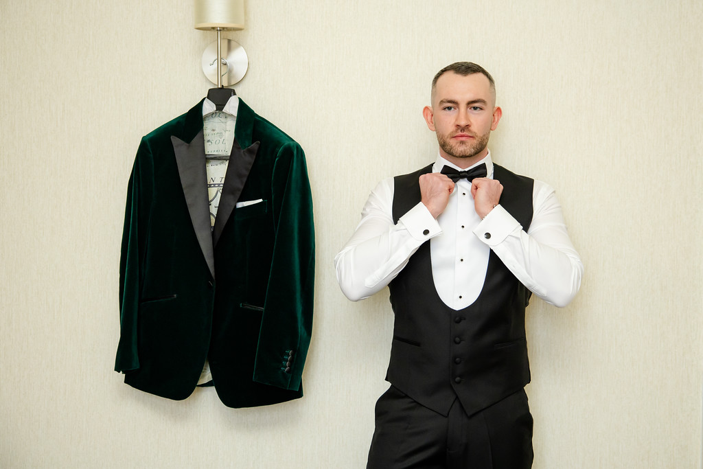 Man in formal attire adjusts his bow tie, standing next to a green velvet jacket hanging on a wall peg. Copyright Somerside Photography Ltd // www.somersidephotography.com
