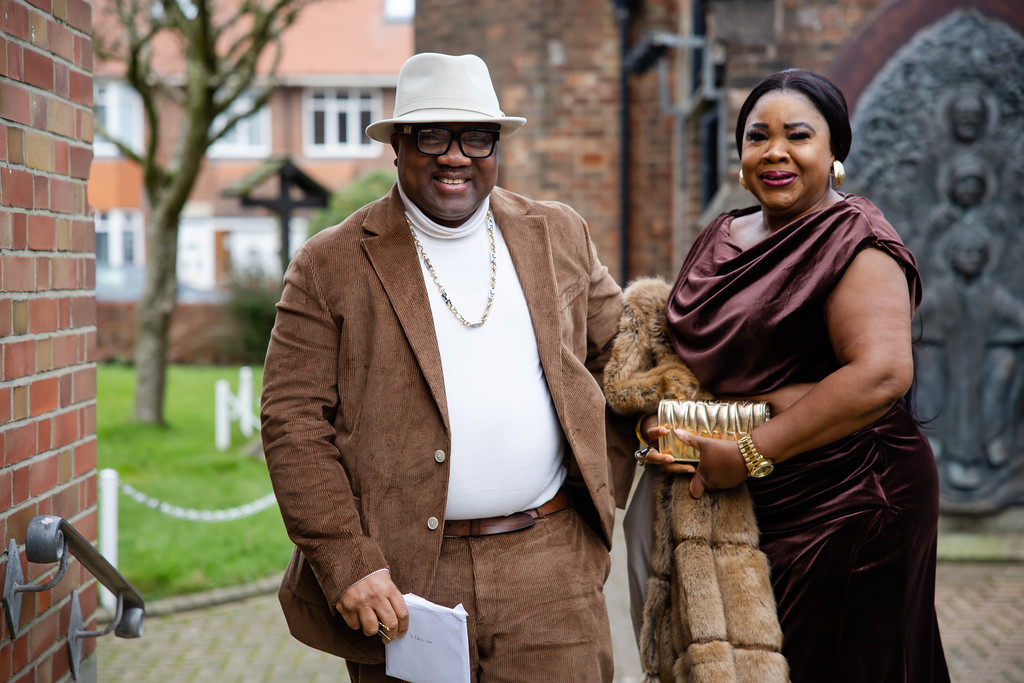 A man in a brown suit and hat stands next to a woman in a brown dress and fur stole, both smiling outdoors near brick buildings. Copyright Somerside Photography Ltd // www.somersidephotography.com