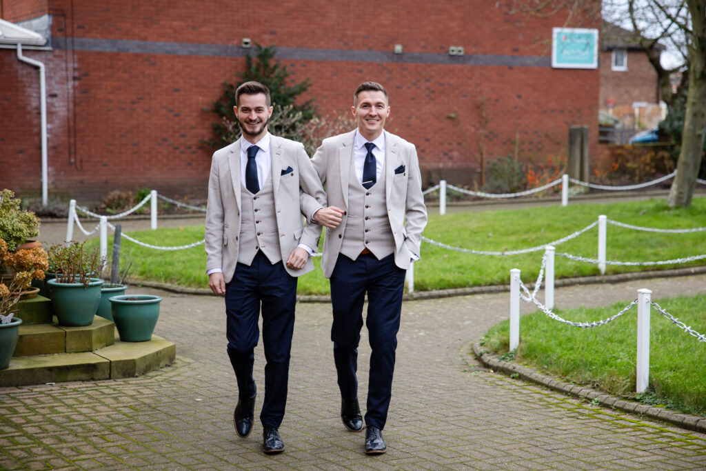 Two men dressed in matching beige jackets, navy trousers, and waistcoats walk arm in arm on a paved path outside a brick building. Copyright Somerside Photography Ltd // www.somersidephotography.com