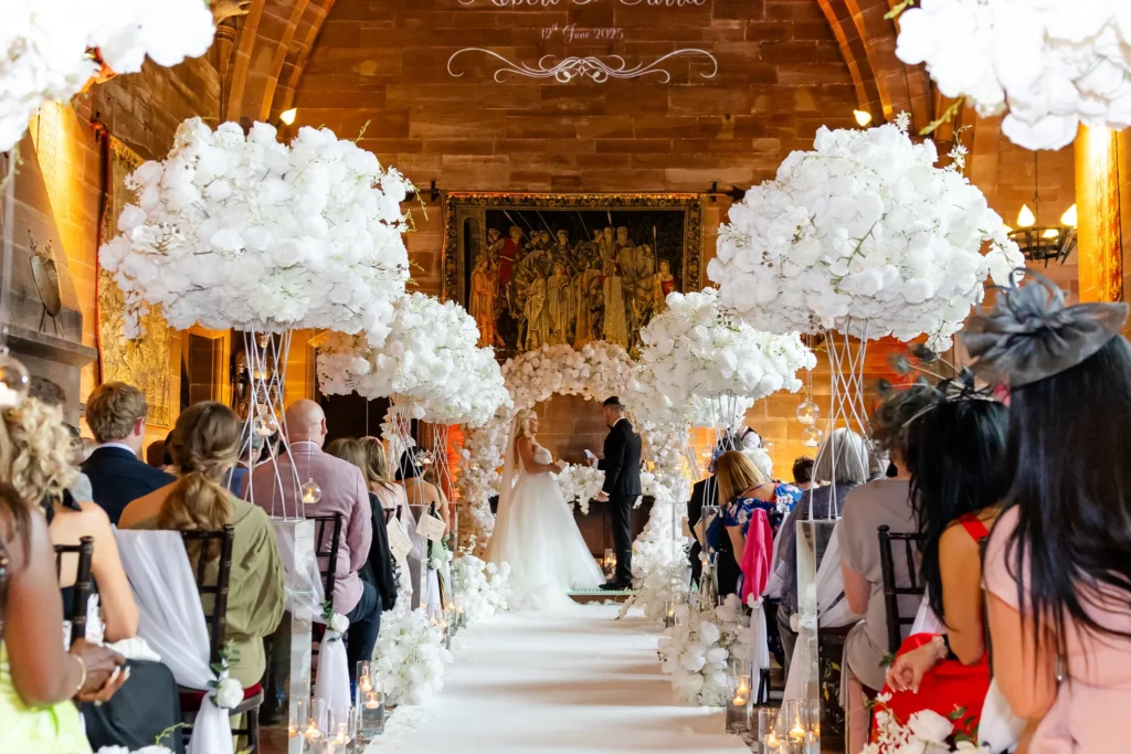 A bride and groom stand at the altar during a wedding ceremony in a decorated hall with white floral arrangements and guests seated on both sides of the aisle. Copyright Somerside Photography Ltd // www.somersidephotography.com