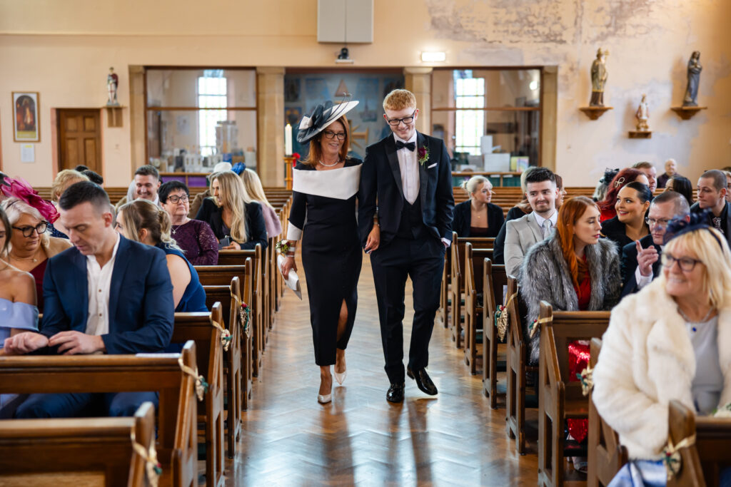 A well-dressed woman and young man walk down the aisle of a church, smiling, whilst seated guests watch and smile during a formal event. Copyright Somerside Photography Ltd // www.somersidephotography.com