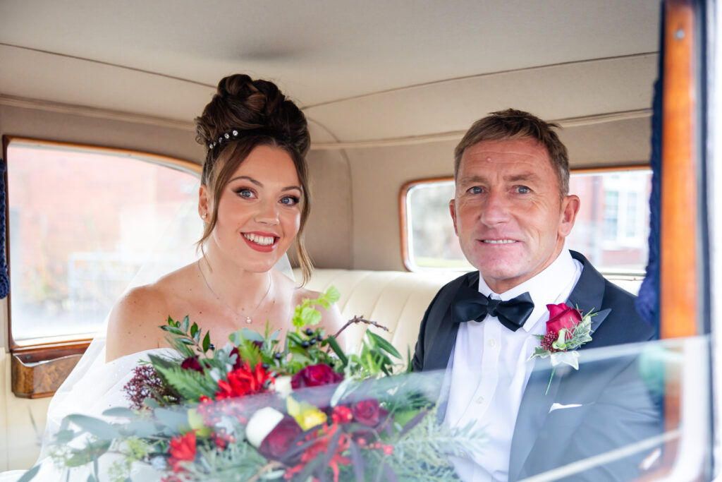 A bride in a white dress and a man in a tuxedo sit together in the back seat of a car, both smiling and holding a bunch of red and green flowers. Copyright Somerside Photography Ltd // www.somersidephotography.com