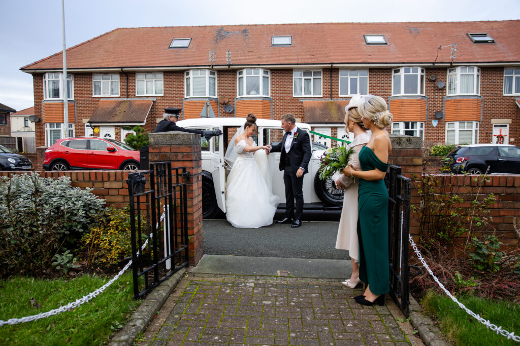 A bride and groom stand beside a vintage white car outside a residential house, with two bridesmaids nearby; a chauffeur waits by the car door. Copyright Somerside Photography Ltd // www.somersidephotography.com