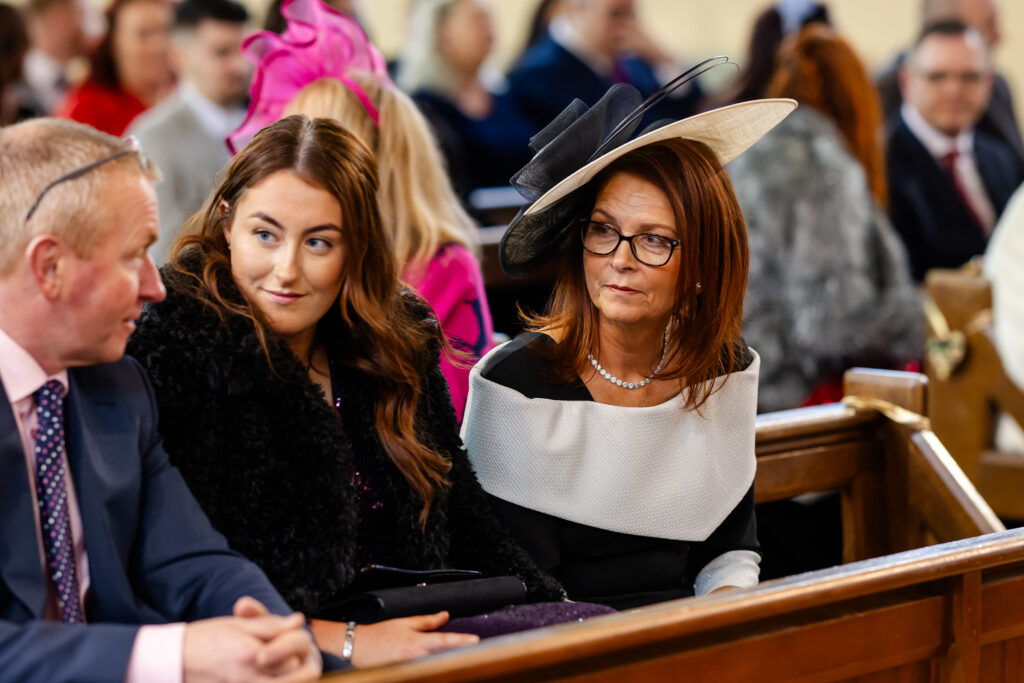 Two women and a man sit in a church pew dressed formally, with the women wearing decorative hats and the man in a suit, surrounded by other seated people. Copyright Somerside Photography Ltd // www.somersidephotography.com
