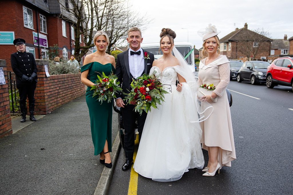 A bride in a white dress stands with a man in a tuxedo, a woman in a green dress, and a woman in a light pink outfit, posing on a street with bouquets of flowers. Copyright Somerside Photography Ltd // www.somersidephotography.com