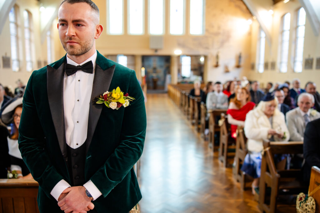 A man in a green velvet suit with a bow tie stands at the front of a church aisle, with seated guests visible in the background. Copyright Somerside Photography Ltd // www.somersidephotography.com