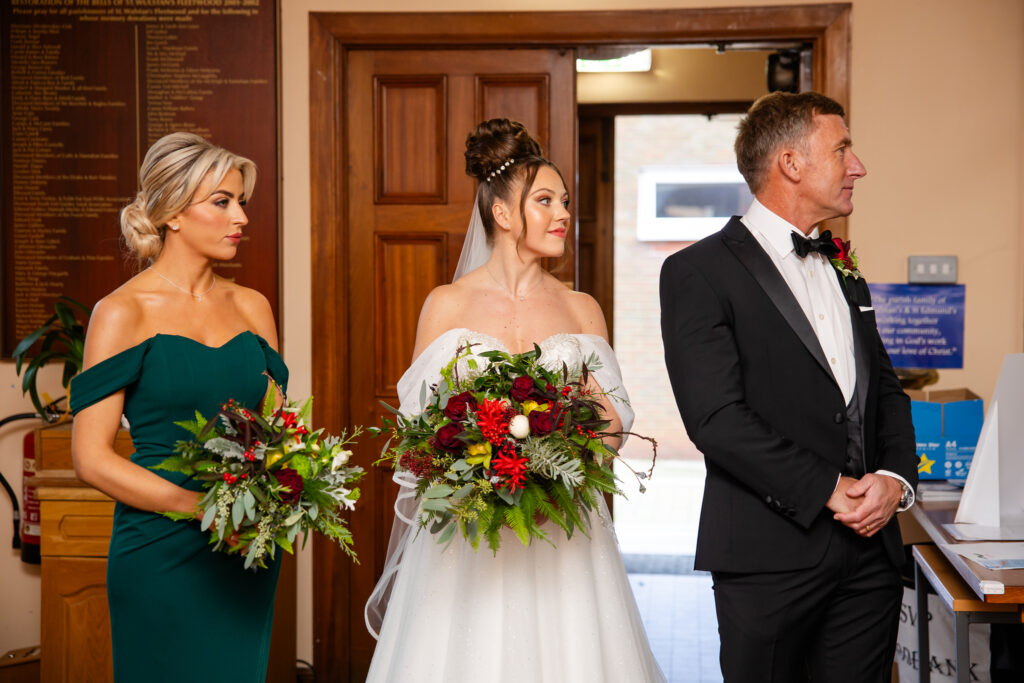 A bride in a white gown stands between a bridesmaid in a green dress and a man in a dinner suit; all are holding bouquets and facing right indoors. Copyright Somerside Photography Ltd // www.somersidephotography.com