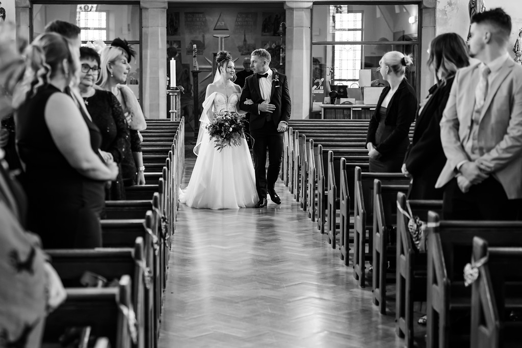 A bride in a wedding dress walks down the aisle with a man as guests watch from pews inside a church. Copyright Somerside Photography Ltd // www.somersidephotography.com