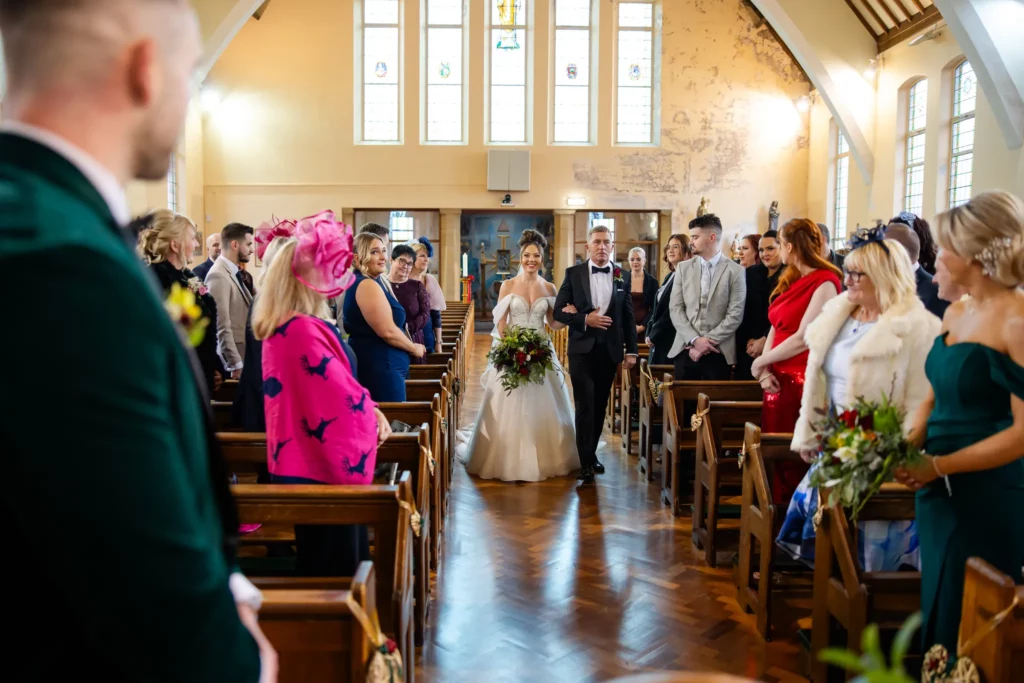 A bride in a white dress walks down the aisle with an older man in a church, as guests on both sides watch and smile. Copyright Somerside Photography Ltd // www.somersidephotography.com