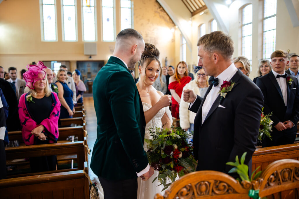 A bride smiles as she shares a fist bump with an older man, while a groom stands beside her in a church filled with seated guests. Copyright Somerside Photography Ltd // www.somersidephotography.com
