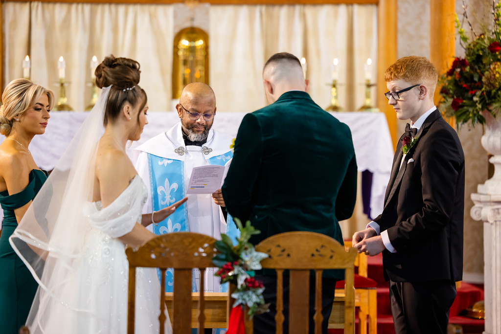A wedding ceremony in a church with a priest reading vows to a bride and groom, joined by two attendants, standing at the altar. Copyright Somerside Photography Ltd // www.somersidephotography.com