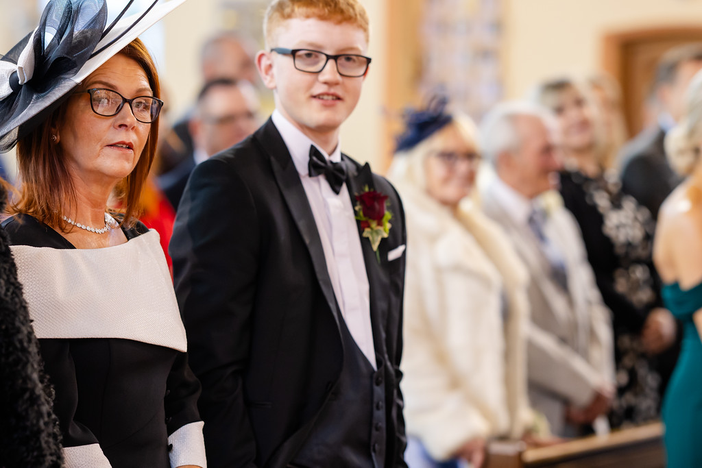 A woman in a black and white dress and large hat stands next to a young man in a dinner jacket at an indoor formal event, with people seated in the background. Copyright Somerside Photography Ltd // www.somersidephotography.com