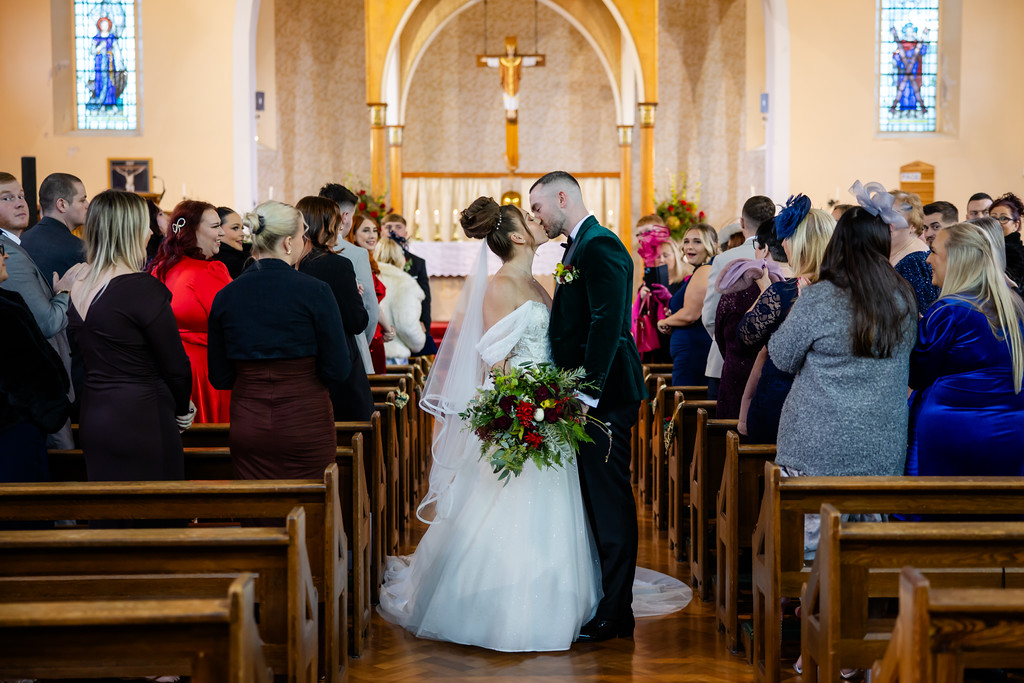 A bride and groom stand at the altar in a church, kissing whilst guests seated in pews watch and take photos. Copyright Somerside Photography Ltd // www.somersidephotography.com