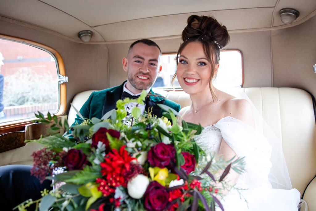 A bride and groom sit together in the back seat of a car, smiling at the camera with a large bouquet of colourful flowers in the foreground. Copyright Somerside Photography Ltd // www.somersidephotography.com