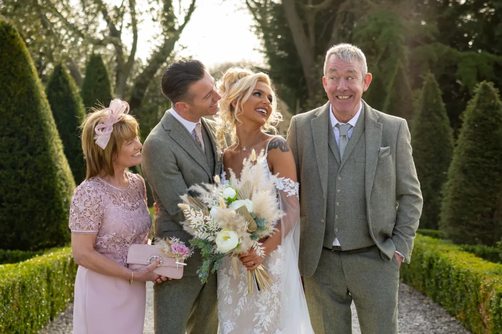 A bride and groom stand outdoors smiling with two older adults, all dressed in formal attire, surrounded by greenery. Copyright Somerside Photography Ltd // www.somersidephotography.com