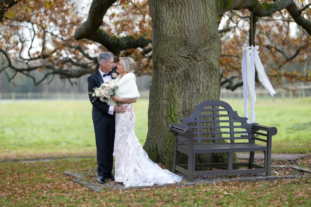 A bride and groom stand together in front of a large tree and a bench outdoors, holding a bouquet, dressed in formal wedding attire. Copyright Somerside Photography Ltd // www.somersidephotography.com