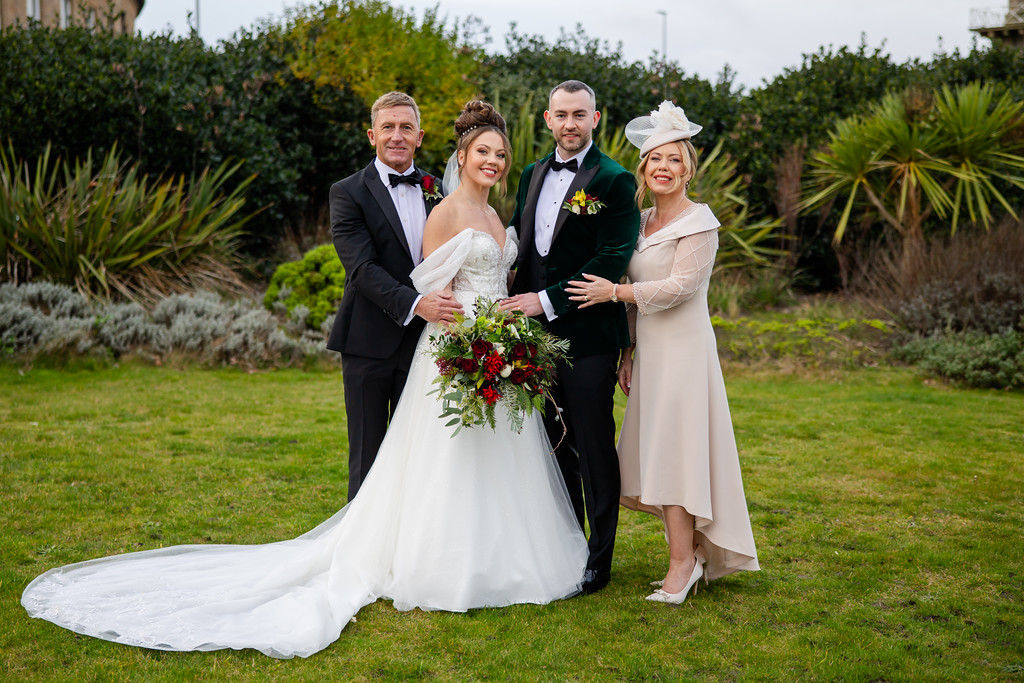 Four adults, two men in dinner jackets and two women in formal dresses, stand together on a lawn with greenery behind them. The bride holds a bouquet and wears a white dress. Copyright Somerside Photography Ltd // www.somersidephotography.com