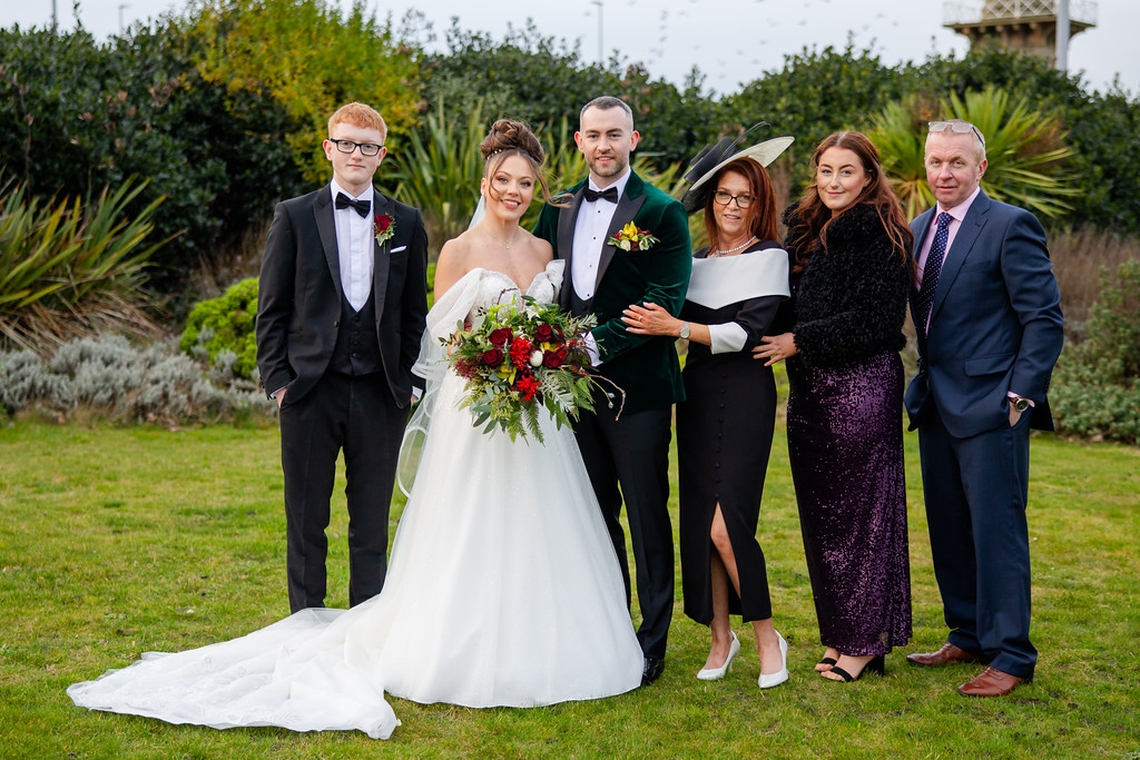 Six people pose outdoors for a formal wedding photo; the bride holds a bouquet, and the group is dressed in formal attire, standing on grass with greenery in the background. Copyright Somerside Photography Ltd // www.somersidephotography.com
