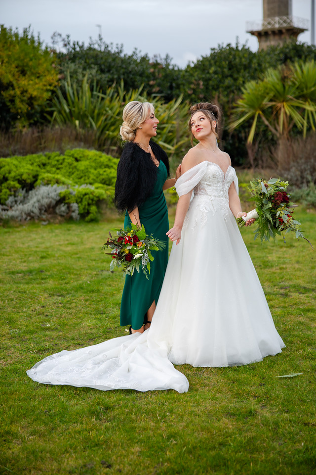 A bride in a white dress stands on grass holding a bouquet, with a bridesmaid in a dark green dress beside her, both looking in different directions. Copyright Somerside Photography Ltd // www.somersidephotography.com
