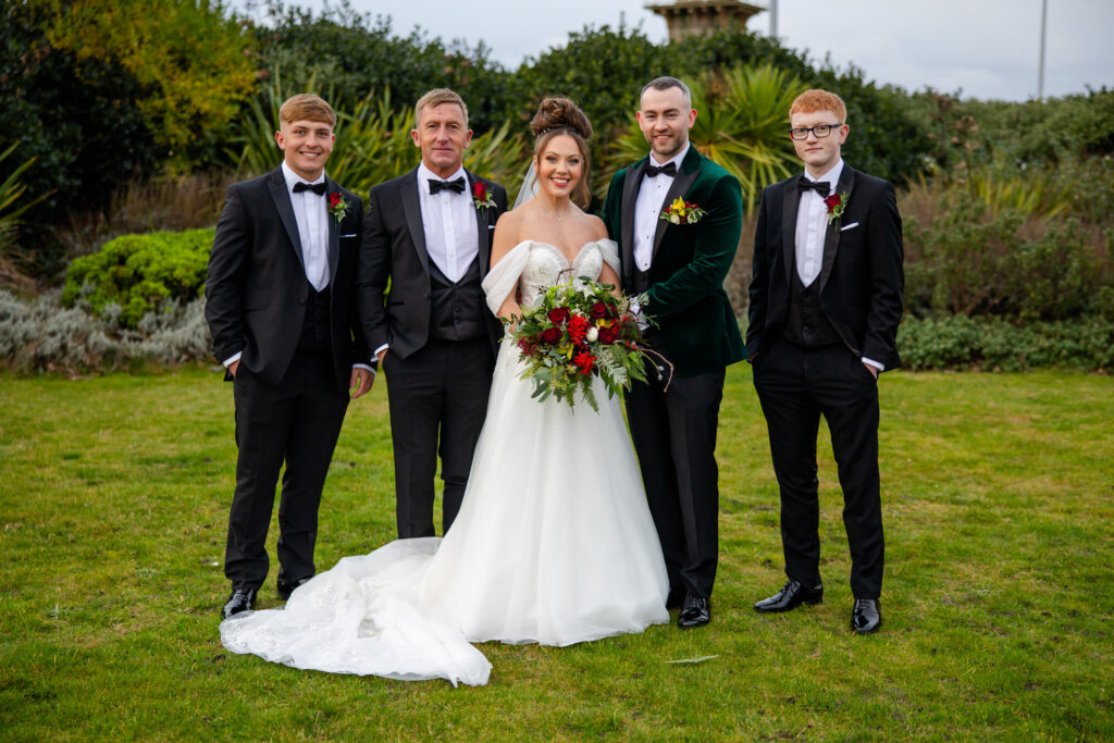 Five people dressed formally pose for a photo outdoors; a bride in a white dress stands in the centre holding a bouquet, flanked by four men in suits. Copyright Somerside Photography Ltd // www.somersidephotography.com