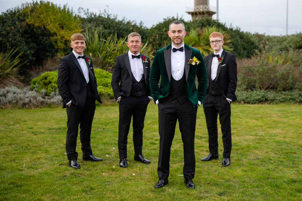Four men in formal suits pose outdoors on grass, with the man in front wearing a green velvet jacket. Bushes and a tall structure are visible in the background. Copyright Somerside Photography Ltd // www.somersidephotography.com
