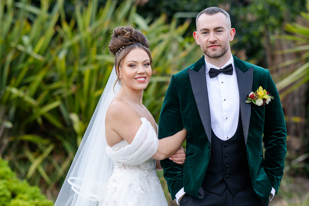 A bride in a white dress and veil stands beside a groom in a dark green velvet jacket, white shirt, and bow tie, posing outdoors in front of greenery. Copyright Somerside Photography Ltd // www.somersidephotography.com