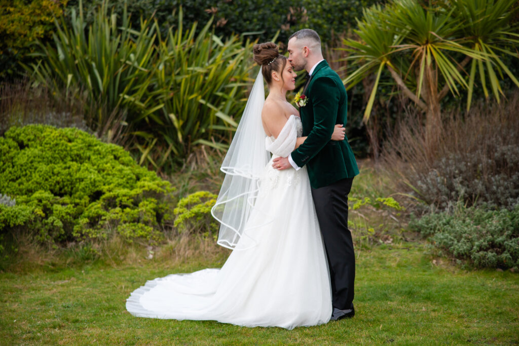 A bride and groom embrace and kiss outdoors in a garden, surrounded by greenery and plants. Copyright Somerside Photography Ltd // www.somersidephotography.com
