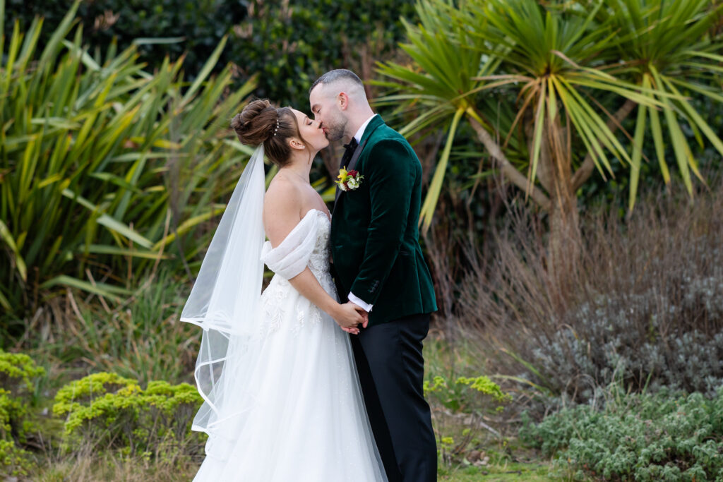 A bride and groom, dressed in wedding attire, kiss outdoors whilst holding hands. They are surrounded by greenery including palm-like plants and shrubs. Copyright Somerside Photography Ltd // www.somersidephotography.com