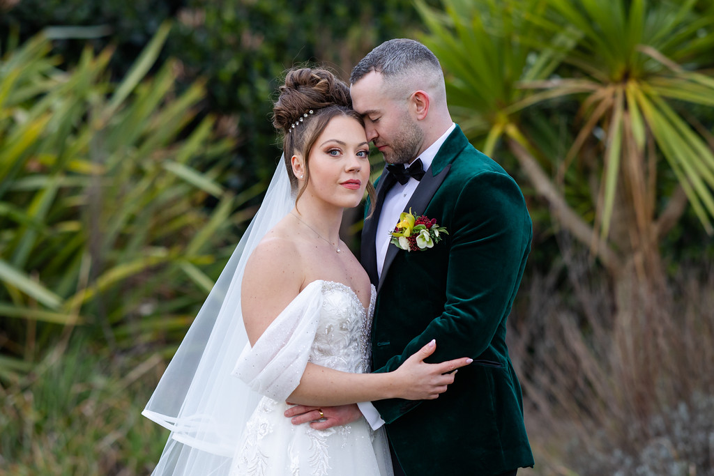 A bride in a white dress and veil stands with a groom in a green velvet suit jacket, embracing outdoors among green foliage. Copyright Somerside Photography Ltd // www.somersidephotography.com