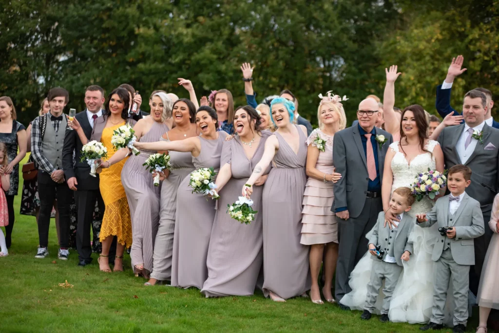 A group of people, including bridesmaids in light purple dresses, groomsmen, and children in suits, pose and smile together outdoors at a wedding. Copyright Somerside Photography Ltd // www.somersidephotography.com